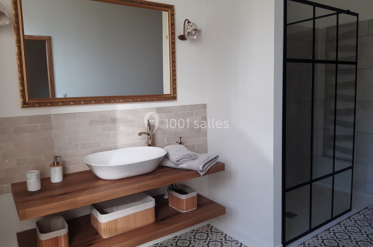 Salle de bain avec lavabo sur meuble en bois, miroir encadré doré, douche à paroi vitrée noire et sol à motifs.