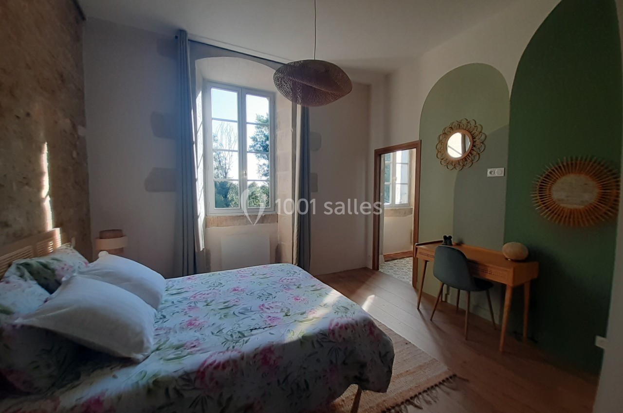 Chambre lumineuse avec lit double, bureau en bois, miroir rond et mur partiellement vert, vue sur jardin par la fenêtre.