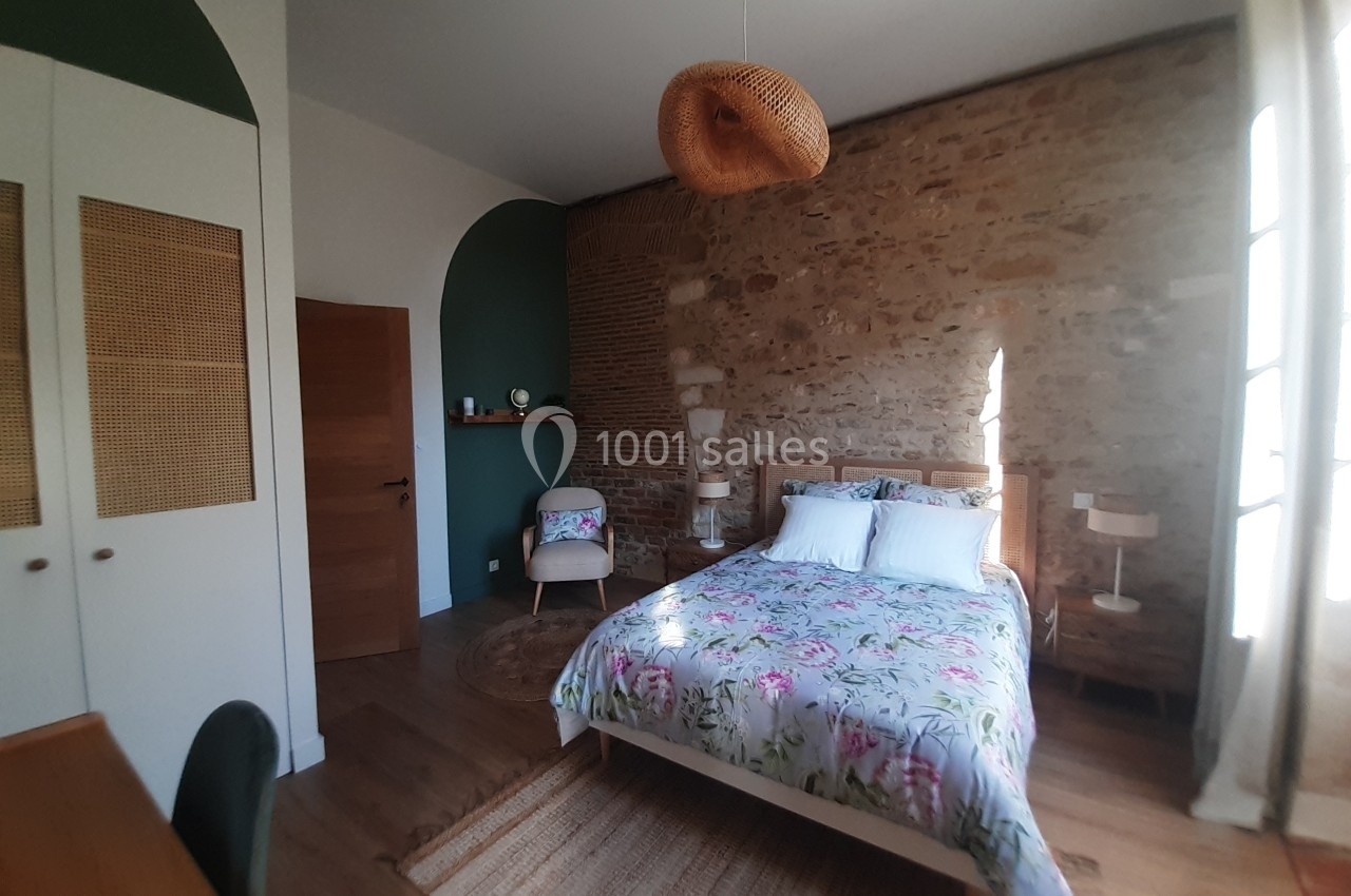 Chambre lumineuse avec mur en pierre, lit double, bureau en bois, fauteuil et suspension en osier.