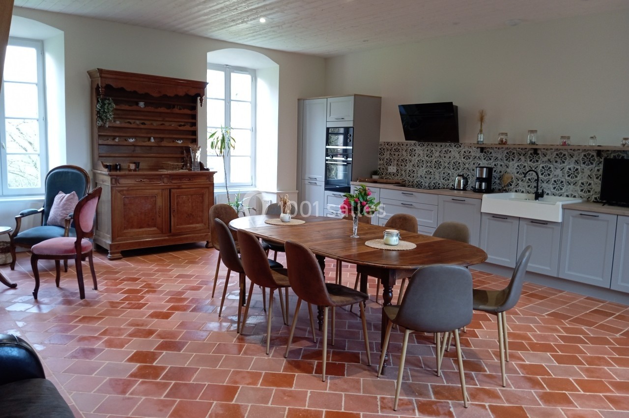 Salle à manger lumineuse avec table en bois, chaises modernes, cuisine équipée et sol en carreaux de terre cuite.