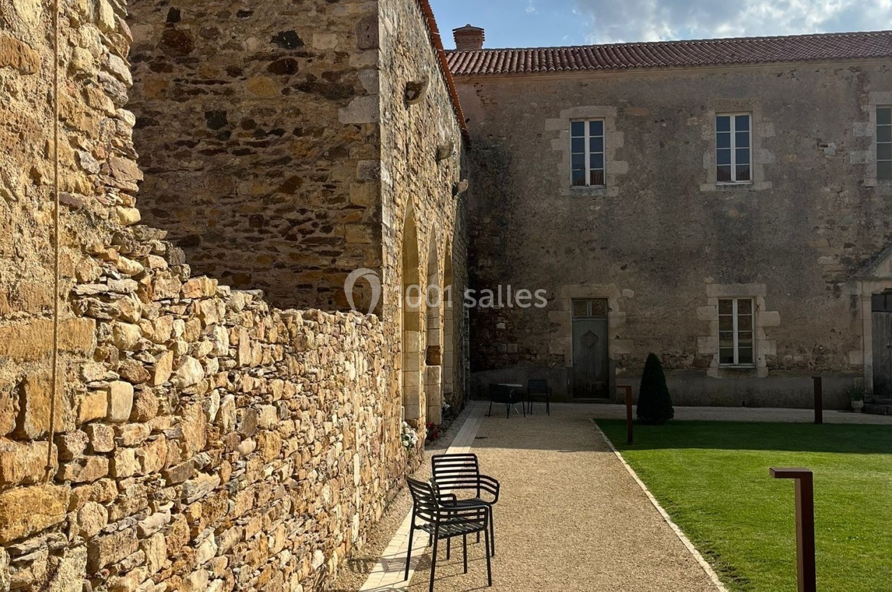 Cour d'un bâtiment en pierre avec bancs métalliques, pelouse et ciel partiellement nuageux en arrière-plan.