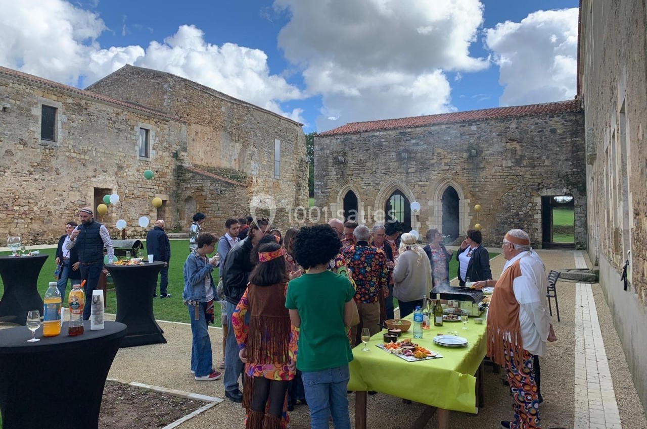 Groupe de personnes rassemblées dans une cour en plein air, près de tables avec nourriture et boissons, sous un ciel nuageux.