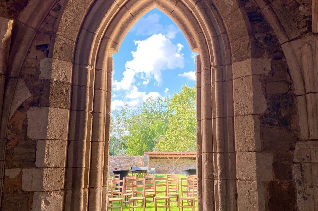 Vue à travers une arche en pierre ancienne donnant sur un jardin ensoleillé avec des chaises en bois et un ciel bleu.