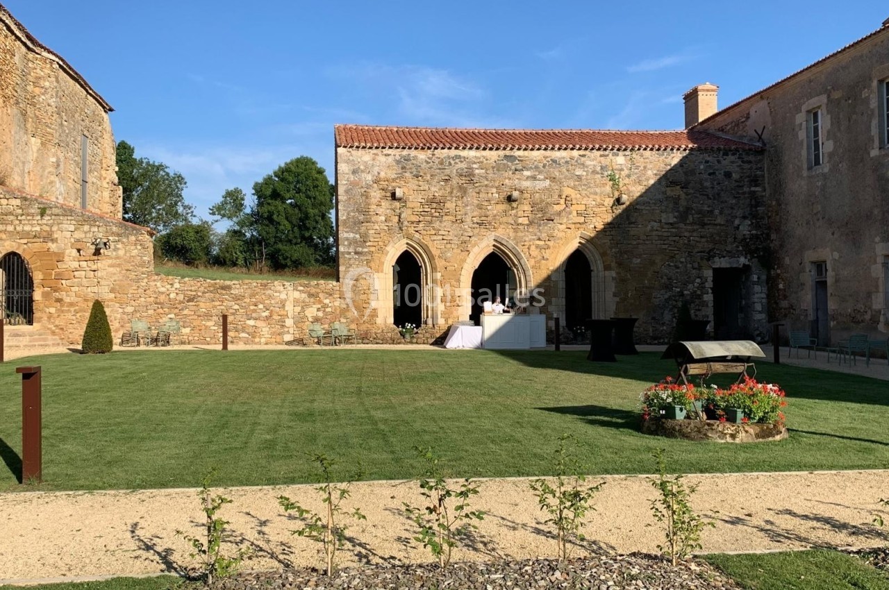 Cour intérieure d'un bâtiment en pierre avec pelouse, parterres fleuris et table dressée sous un ciel dégagé.