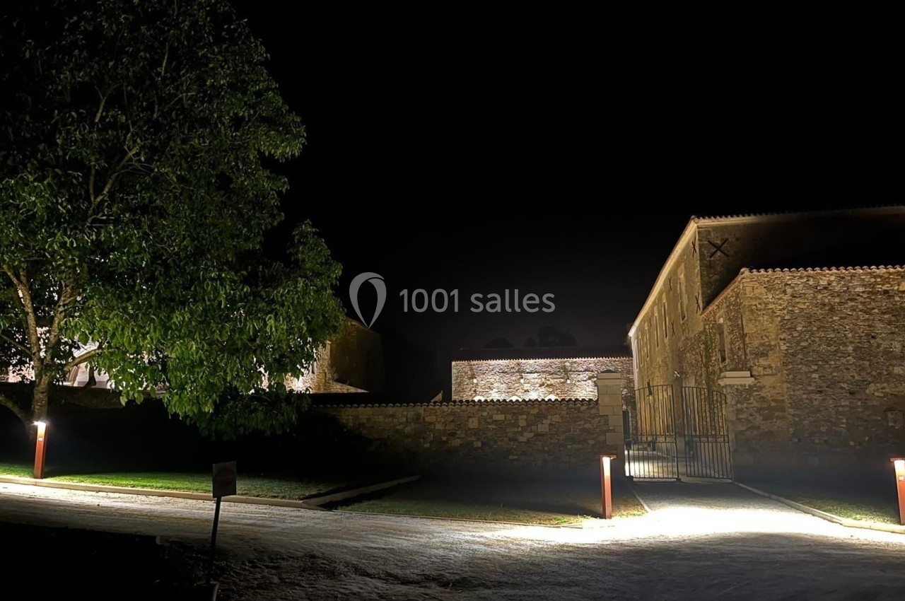 Cour intérieure éclairée la nuit, entourée de bâtiments en pierre et d'un arbre à gauche.