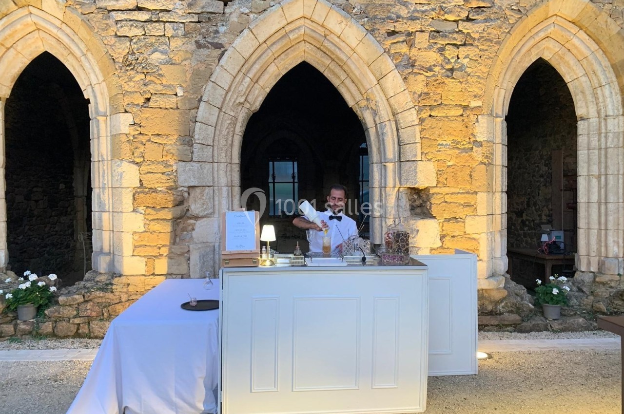 Un serveur prépare une boisson derrière un comptoir blanc installé devant une façade en pierre avec des arches.