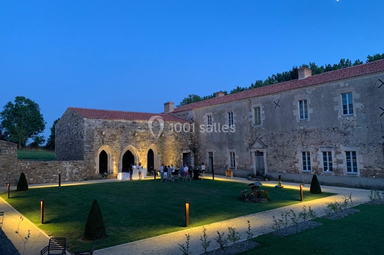 Cour d'un bâtiment historique en pierre, éclairée en soirée avec des arches gothiques et un groupe de personnes rassemblées.