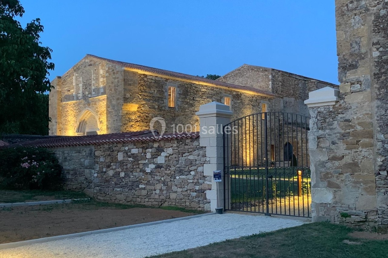 Vue d'un bâtiment en pierre éclairé à la tombée de la nuit, entouré d'un mur et d'une grille ouverte.