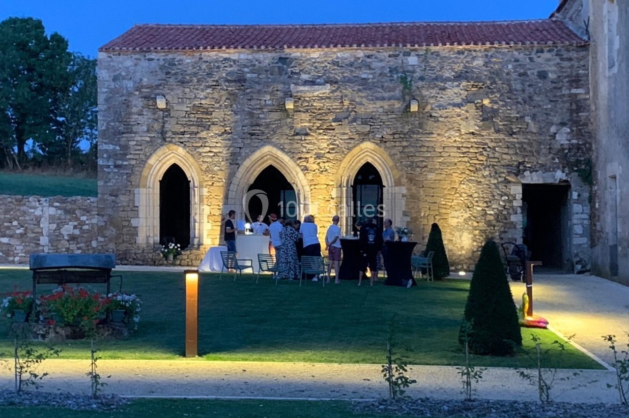 Groupe de personnes rassemblées en soirée devant un bâtiment en pierre éclairé, avec pelouse et décoration lumineuse.