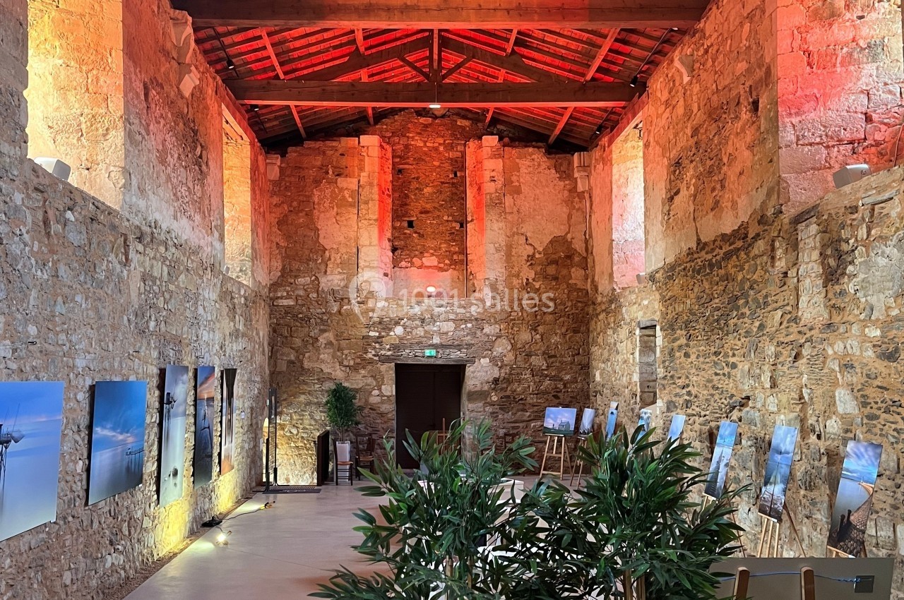 Salle en pierre avec un plafond en bois, éclairée par des lumières rouges et jaunes, exposant des photographies sur des…
