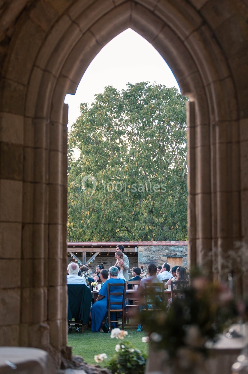 Vue à travers une arche en pierre sur un rassemblement de personnes assises à l'extérieur, près d'un grand arbre.