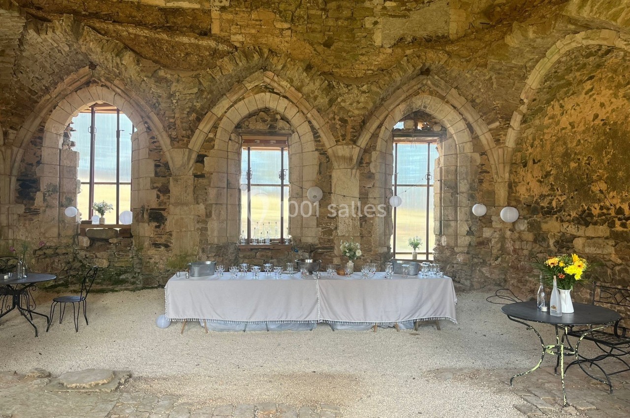 Salle en pierre ancienne avec trois grandes fenêtres gothiques, tables décorées pour un événement festif.