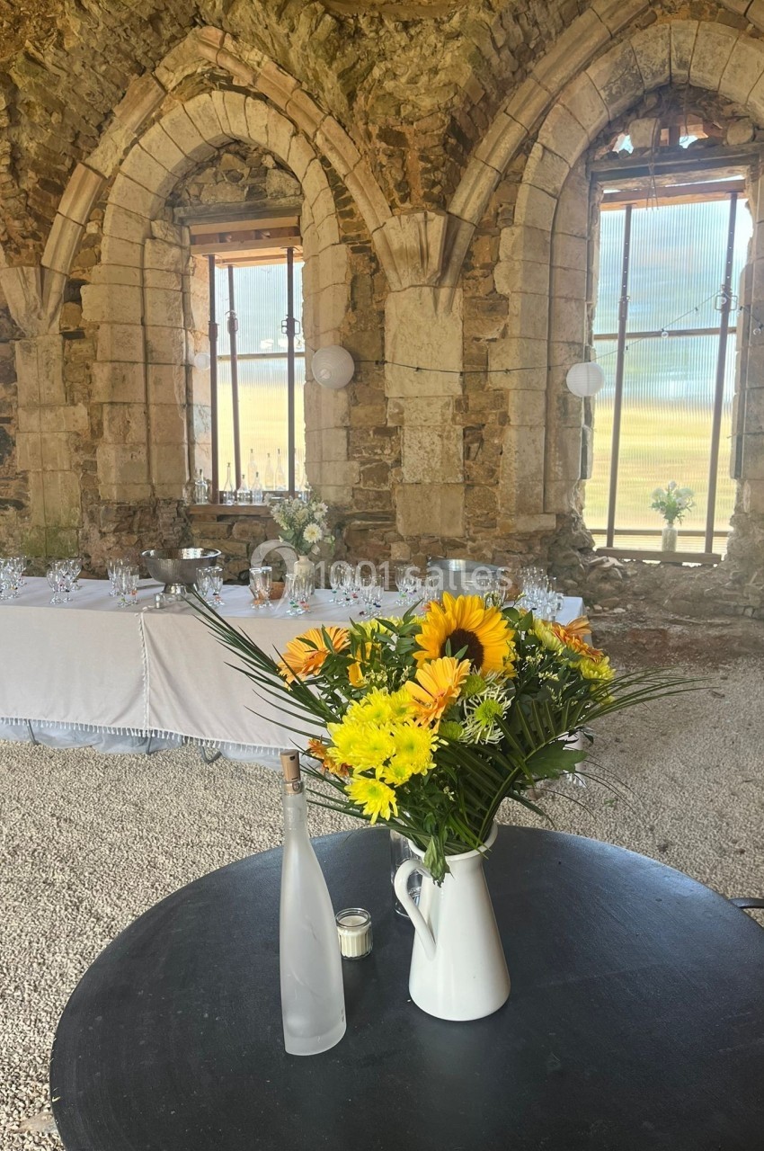 Un bouquet de fleurs jaunes dans un pichet blanc posé sur une table ronde, devant une salle en pierre avec des arches.