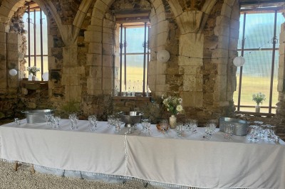Table dressée avec nappes blanches, verres et seaux à glace, dans une salle en pierre avec de grandes fenêtres.