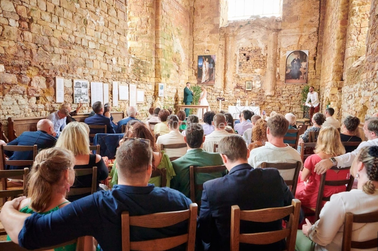 Un prêtre célèbre une messe dans une église en pierre ancienne, avec des fidèles assis sur des chaises en bois.