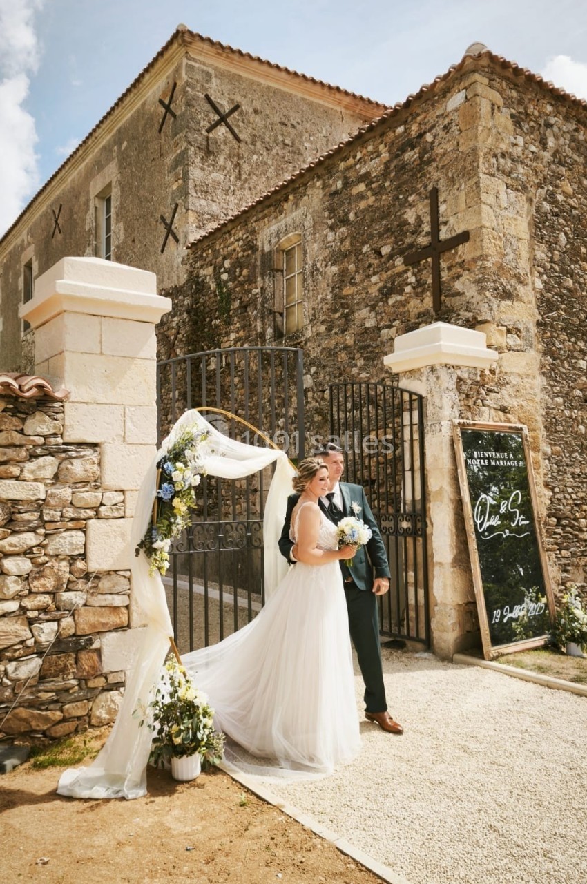 Un couple de mariés pose devant une arche fleurie près d'un bâtiment en pierre avec une porte en fer forgé.