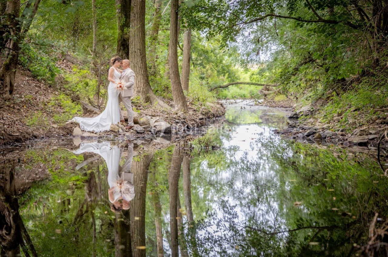 Un couple en tenue de mariage se tient près d'un ruisseau entouré d'arbres, avec leur reflet visible dans l'eau calme.