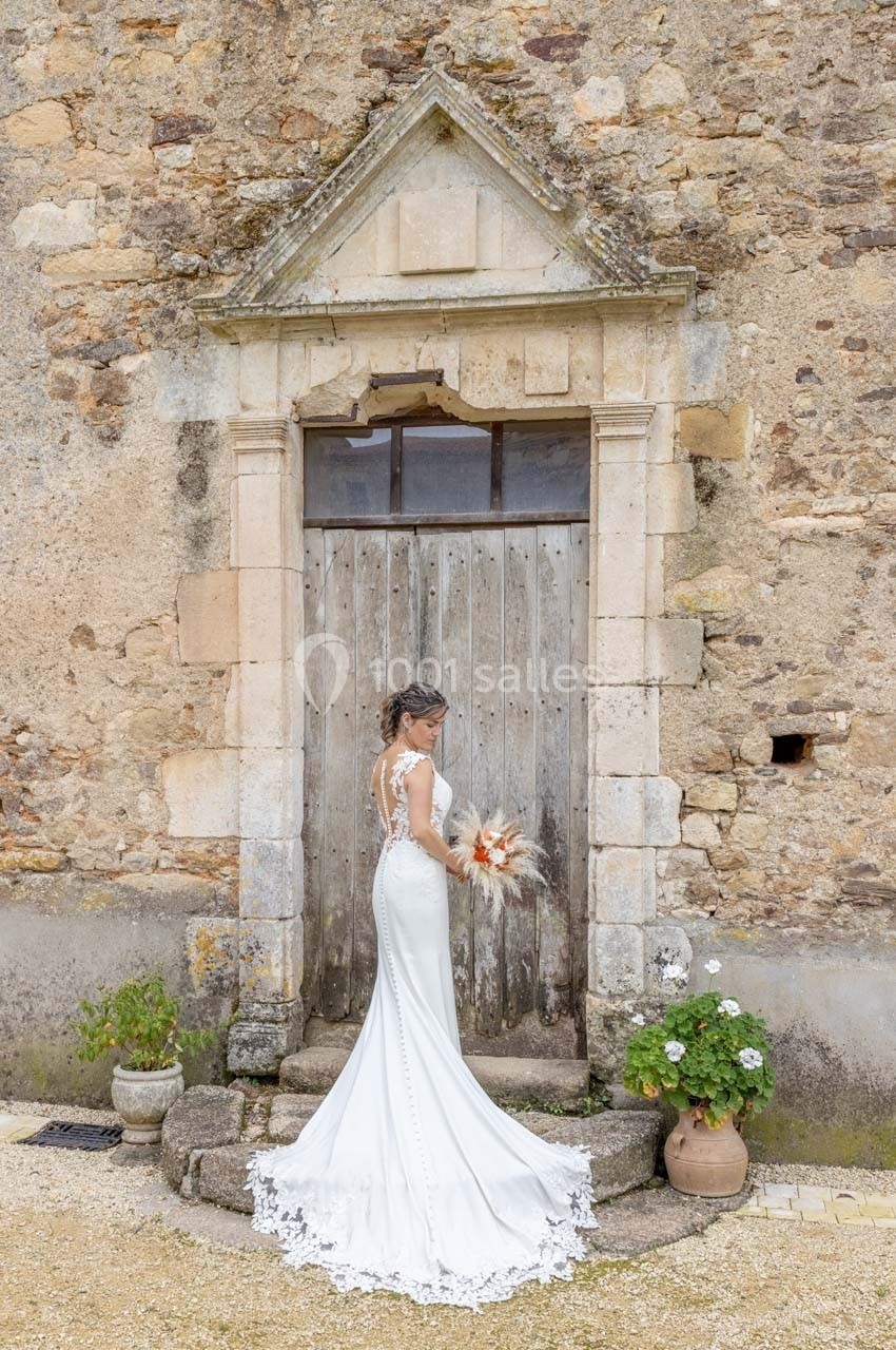 Une mariée en robe blanche tient un bouquet devant une porte en bois encadrée de pierres anciennes.