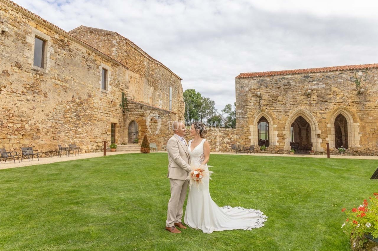 Un couple en tenue de mariage pose dans une cour verdoyante entourée de bâtiments en pierre ancienne.