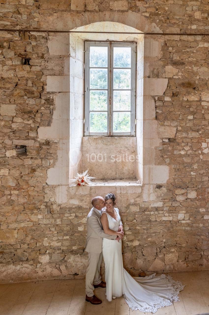 Un couple en tenue de mariage pose devant un mur en pierre avec une grande fenêtre laissant entrer la lumière.
