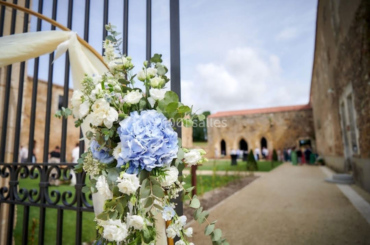 Composition florale avec hortensias bleus et fleurs blanches accrochée à une grille, devant un bâtiment en pierre.