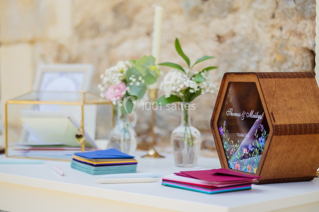 Boîte hexagonale en bois pour cartes, entourée de fleurs, enveloppes colorées et stylos sur une table blanche.