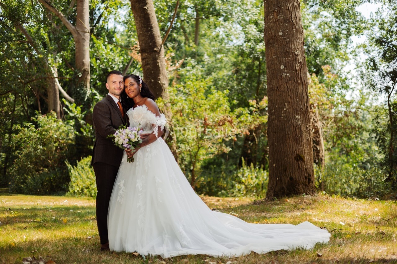 Un couple en tenue de mariage pose dans un parc boisé, entouré de grands arbres et d'une lumière naturelle.