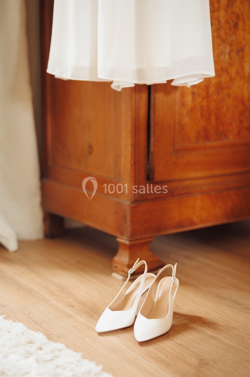 Chaussures blanches à talons posées sur un sol en bois près d'une armoire ancienne et d'un tissu blanc suspendu.