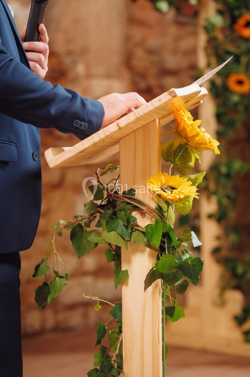 Un orateur en costume tient un micro derrière un pupitre en bois décoré de tournesols et de feuillage.