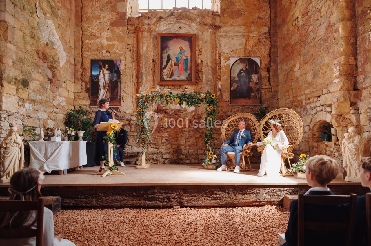 Cérémonie de mariage dans une église en pierre, avec un couple assis sur des fauteuils en osier et un officiant debout.