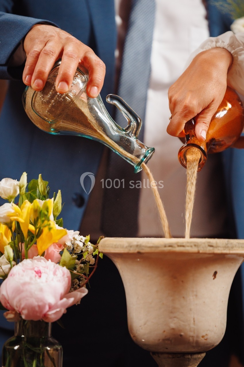 Deux mains versent du sable de deux carafes dans un récipient, entouré de fleurs colorées.