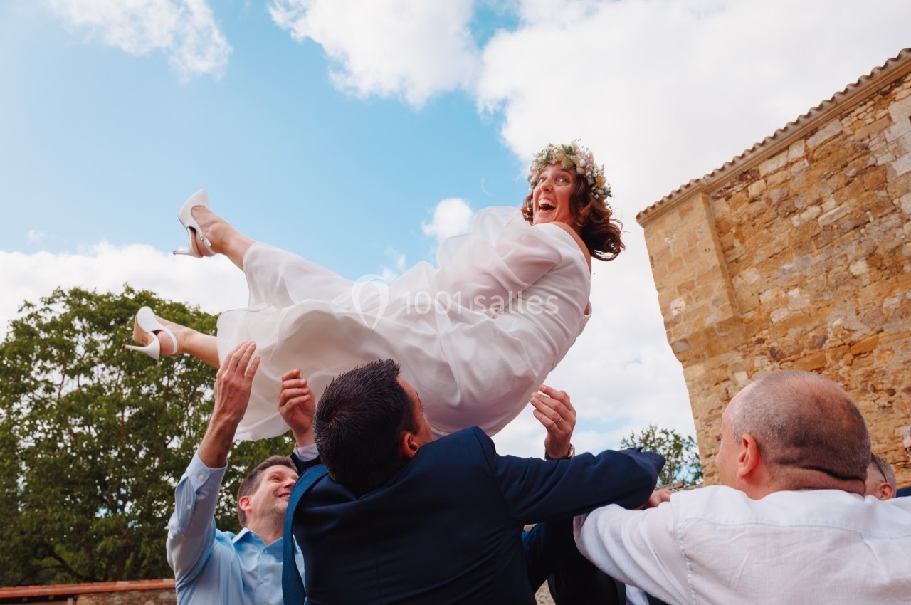 Une mariée souriante en robe blanche est soulevée en l'air par des invités lors d'une célébration en extérieur.