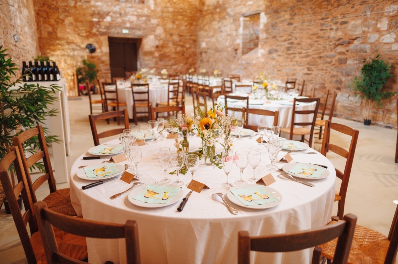 Salle de réception avec tables rondes dressées, décorées de fleurs et de vaisselle élégante, dans un cadre en pierre.