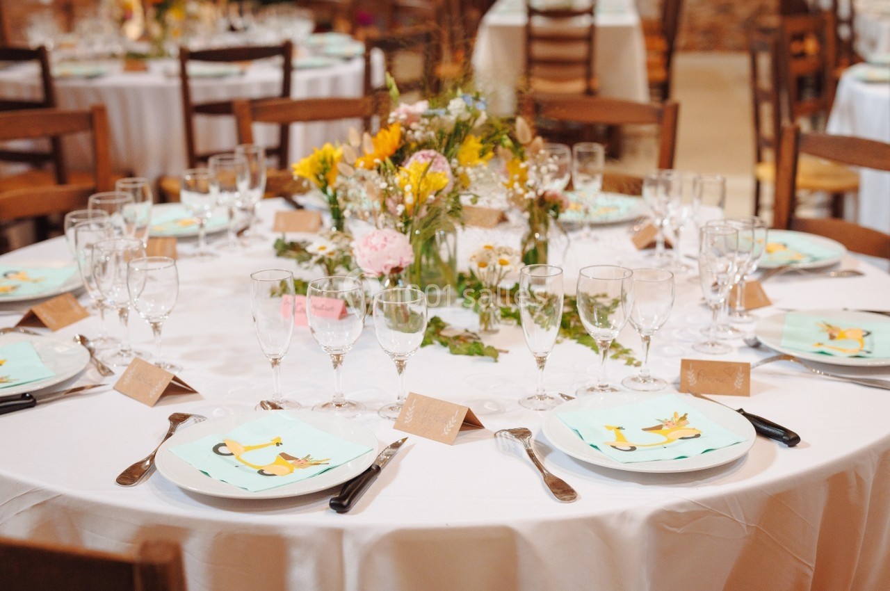 Table dressée pour un événement avec nappes blanches, vaisselle, verres, marque-places et centre de table floral.