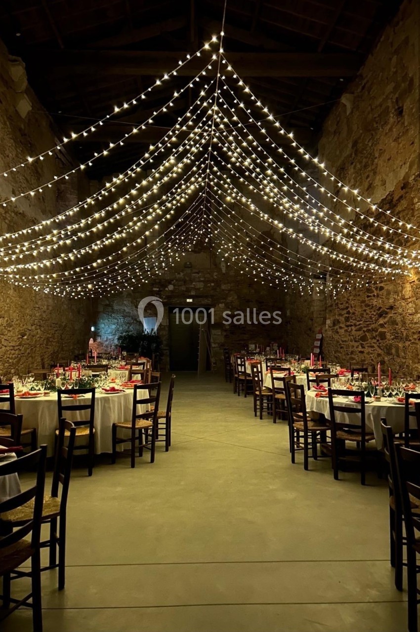 Salle en pierre décorée pour un événement, avec guirlandes lumineuses suspendues et tables dressées.