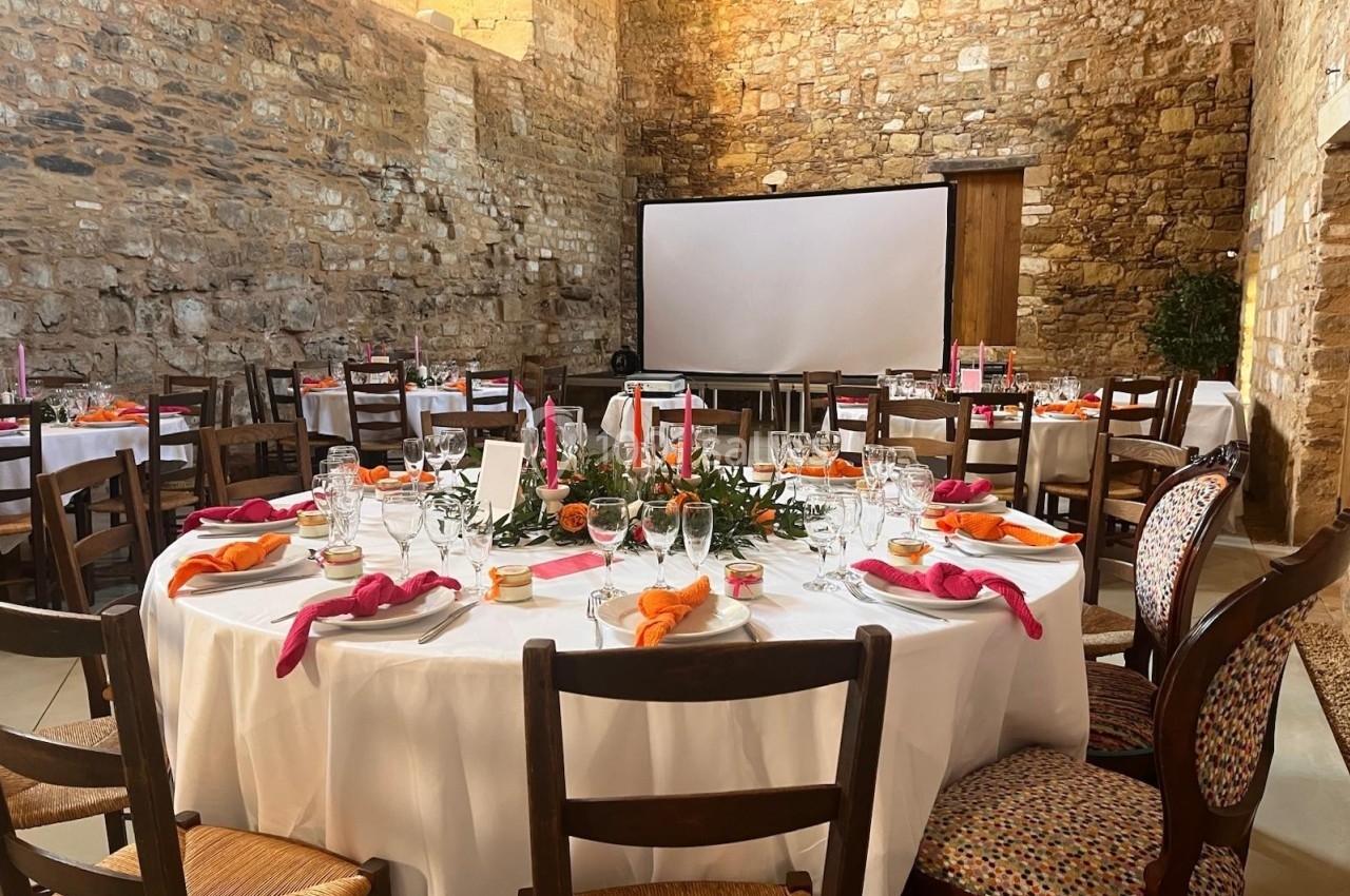 Salle en pierre aménagée pour un événement, avec tables rondes dressées, nappes blanches et décorations colorées.