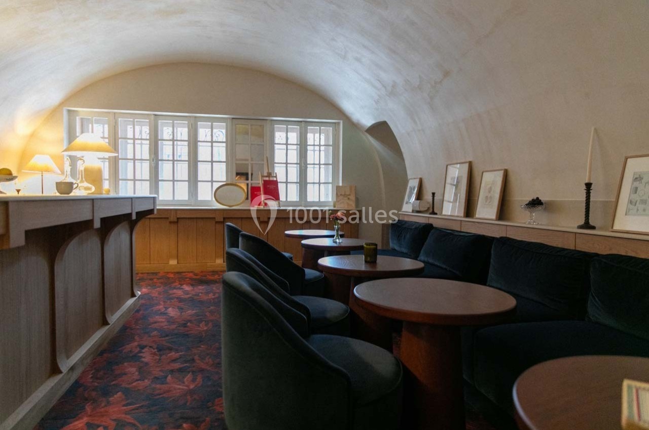 Salon avec canapé en velours, tables rondes en bois, éclairage doux et fenêtres laissant entrer la lumière naturelle.