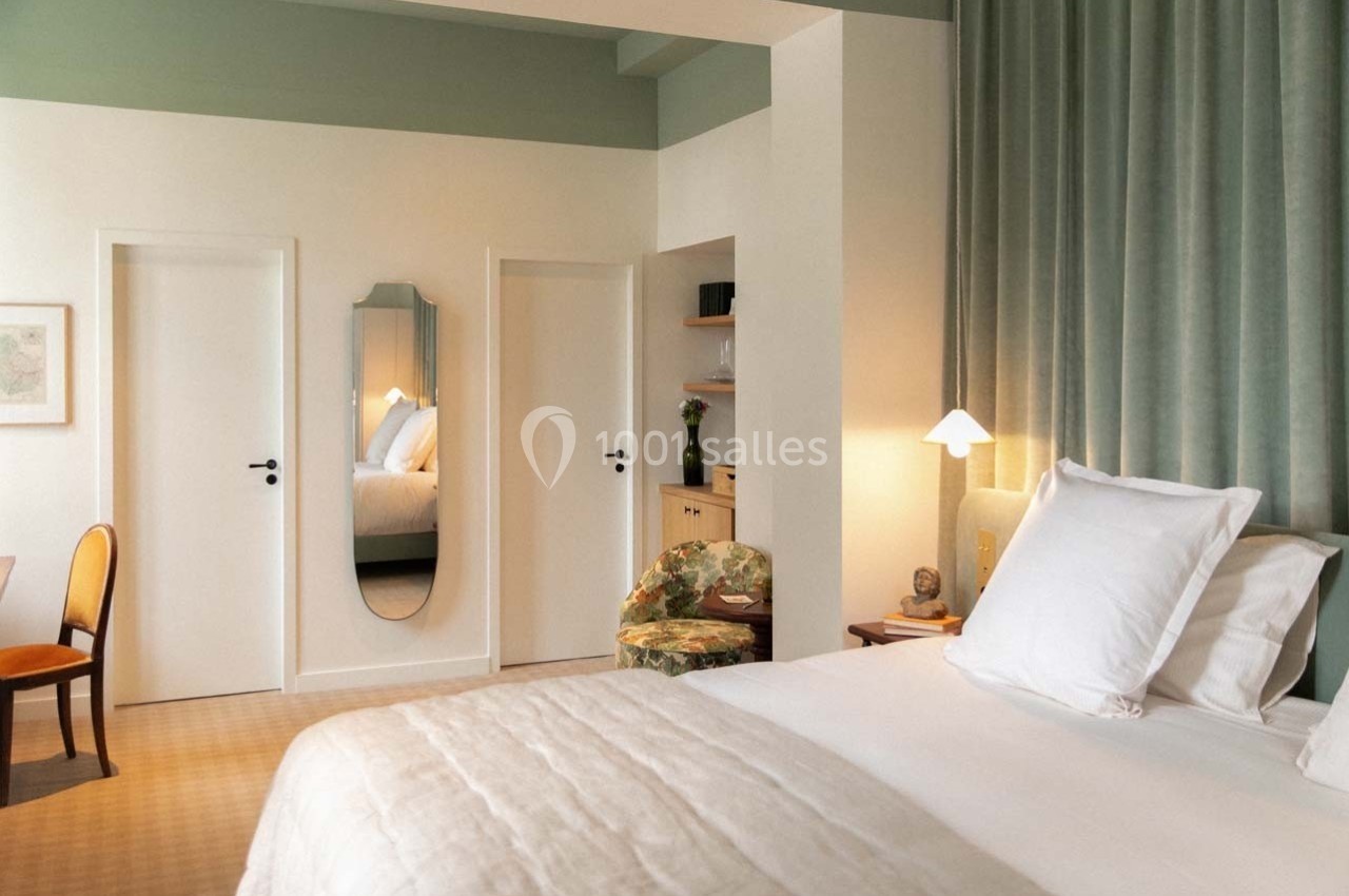 Chambre lumineuse avec lit double, miroir mural, rideaux verts, bureau en bois et chaise rembourrée.