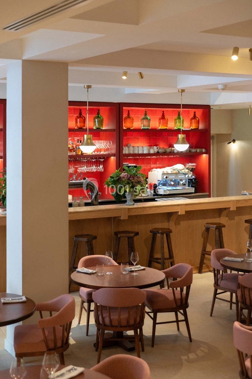 Salle de restaurant avec un bar en bois, étagères rouges décorées de bouteilles, et tables entourées de chaises rembourrées.