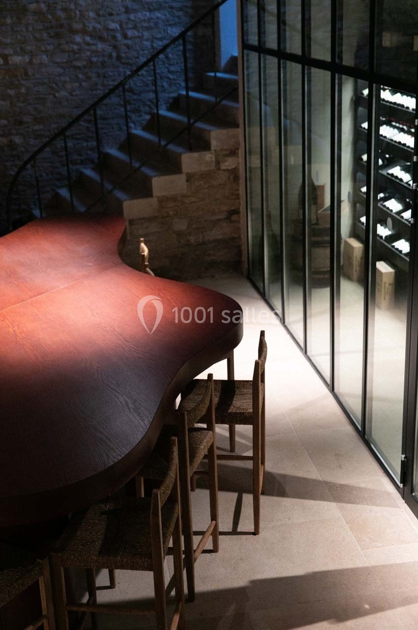 Table en bois aux formes organiques entourée de chaises, près d'une cave vitrée et d'un escalier en pierre.