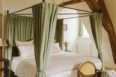 Chambre lumineuse sous combles avec poutres apparentes, lit à baldaquin, table en bois et fenêtre donnant sur l'extérieur.