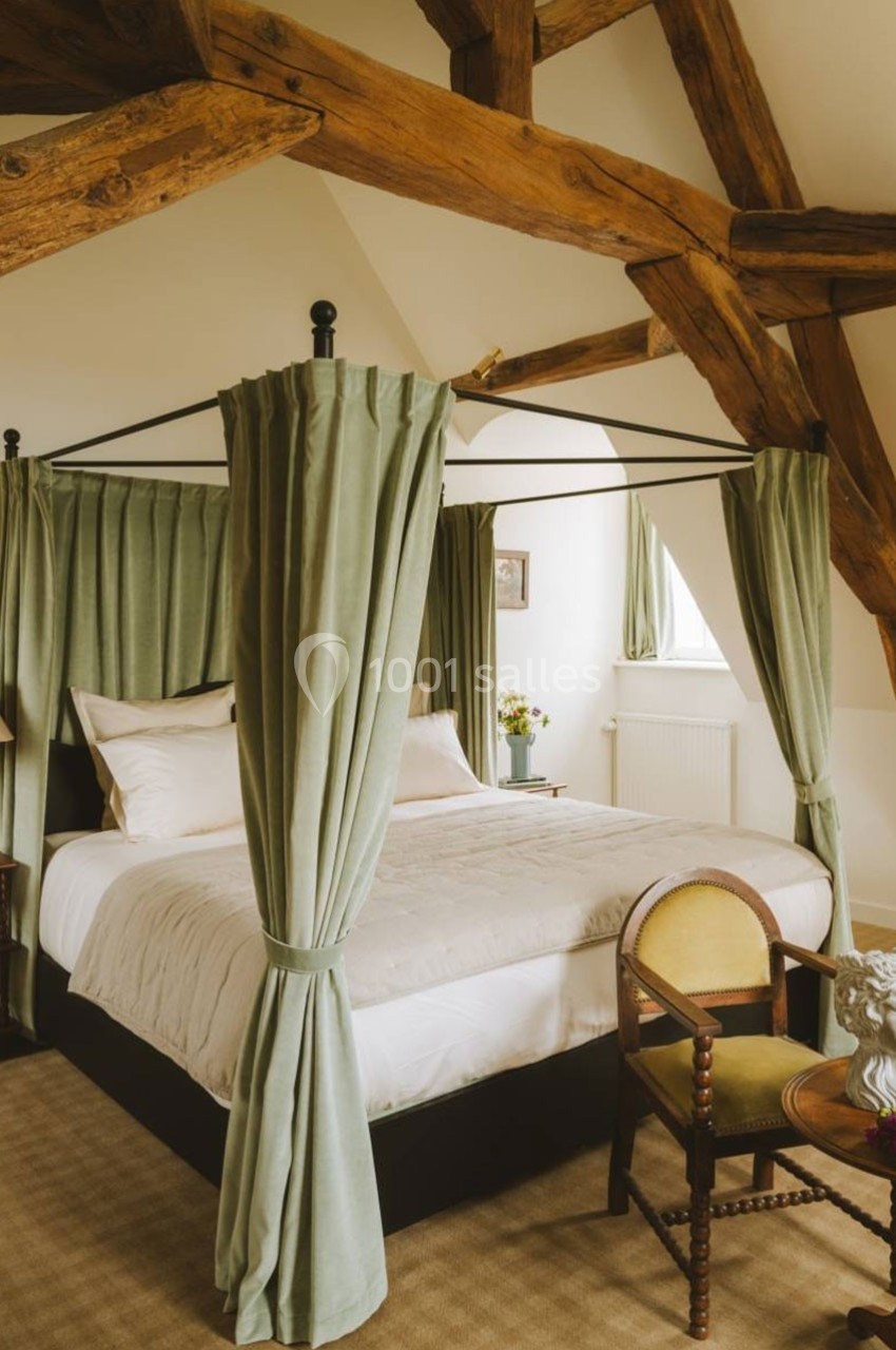 Lit à baldaquin avec rideaux verts dans une chambre lumineuse aux poutres apparentes et mobilier classique.