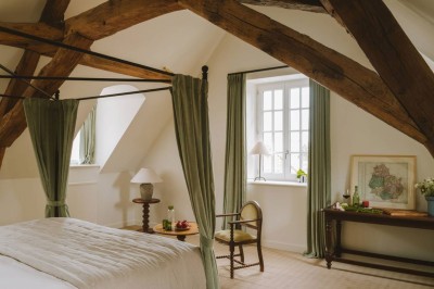 Chambre lumineuse sous combles avec poutres apparentes, lit à baldaquin, table en bois et fenêtre donnant sur l'extérieur.