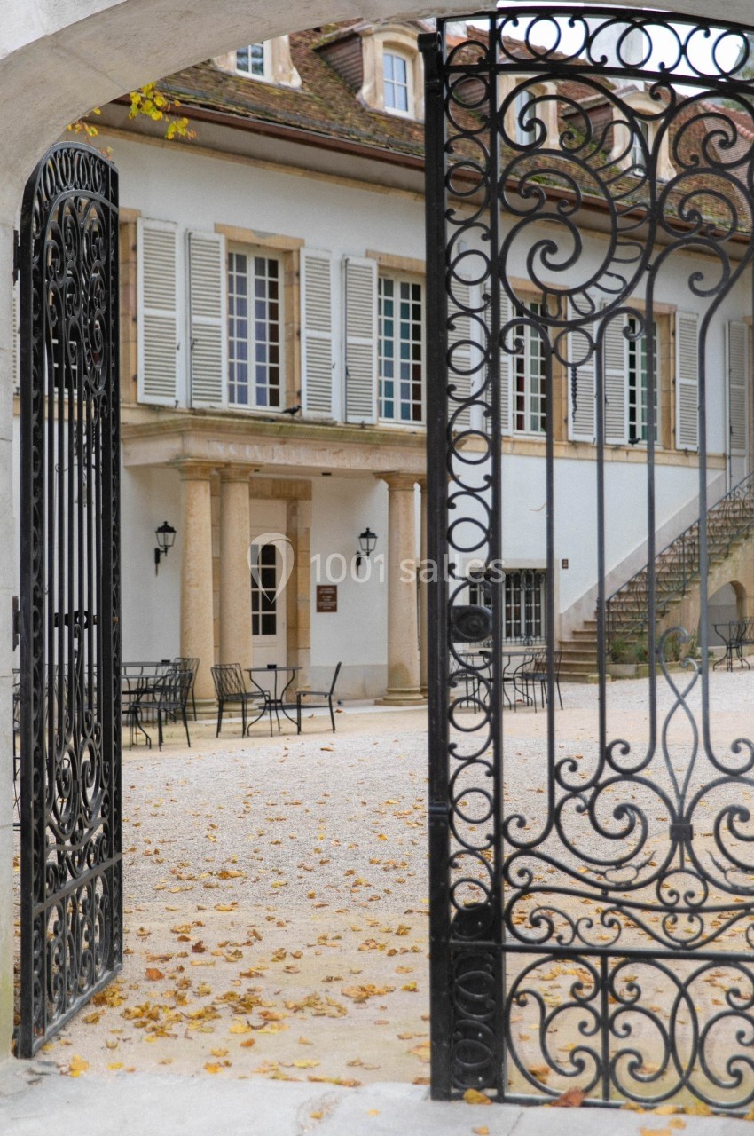 Portail en fer forgé ouvert donnant sur une cour pavée avec des tables, des chaises et une bâtisse aux volets blancs.