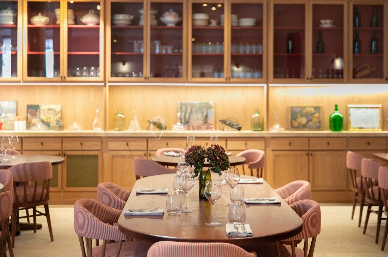 Salle de restaurant lumineuse avec tables dressées, chaises en tissu rose et étagères en bois garnies d'objets décoratifs.
