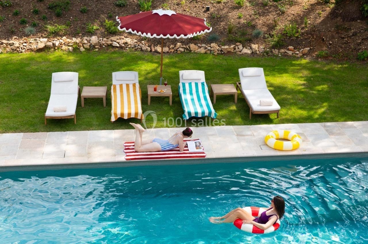 Une femme nage avec une bouée dans une piscine, tandis qu'une autre est allongée sur une serviette près de transats colorés.