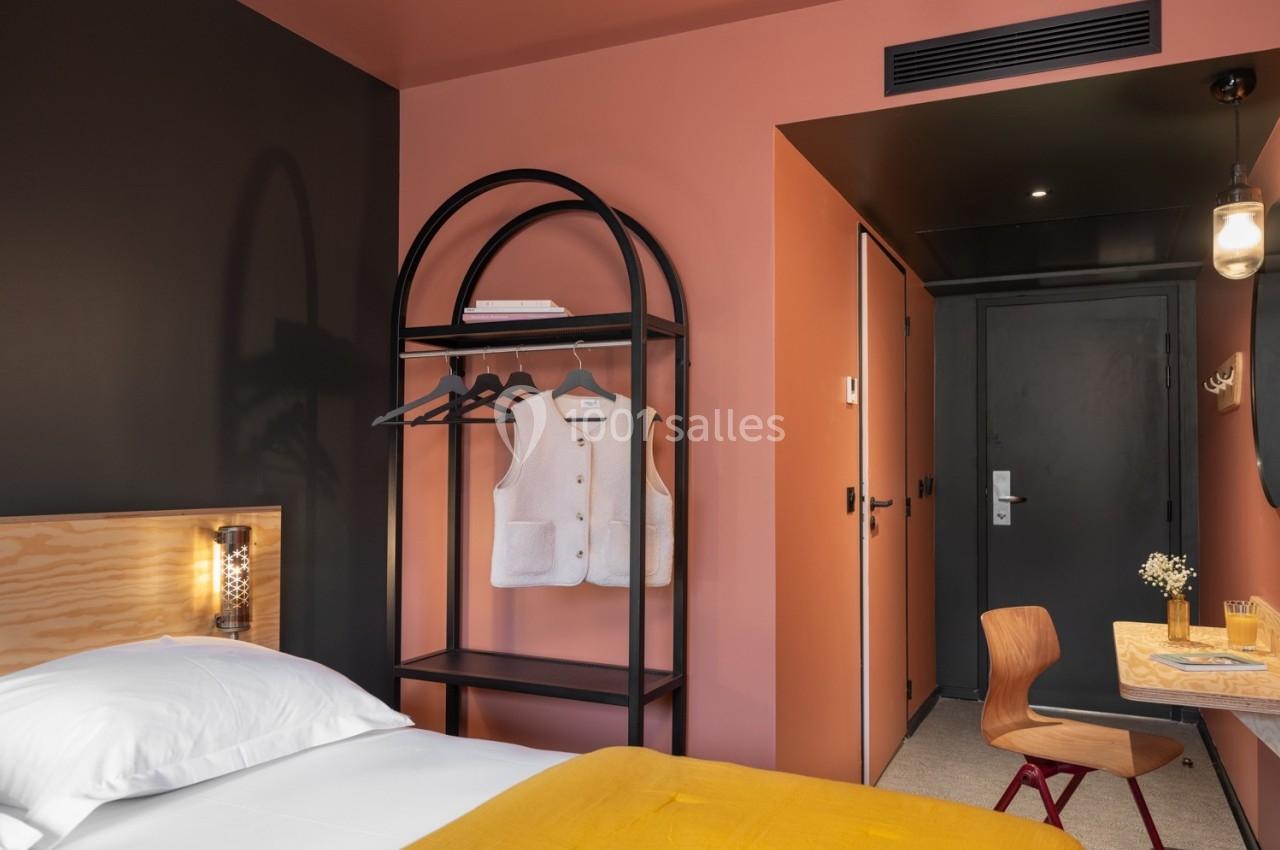 Chambre d'hôtel avec lit, étagère portant des vêtements, bureau en bois et éclairage moderne dans un décor aux tons chauds.