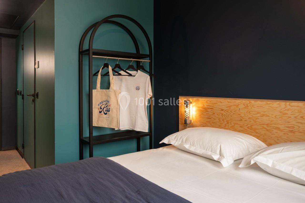 Lit double avec draps blancs et couverture bleue, portant avec vêtements et sac en toile dans une chambre aux murs colorés.