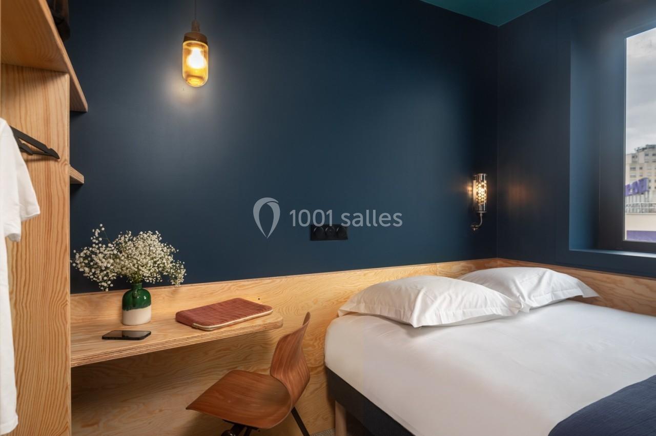 Chambre moderne avec murs bleu foncé, lit simple, bureau en bois, chaise, lampe suspendue et fenêtre donnant sur l'extérieur.