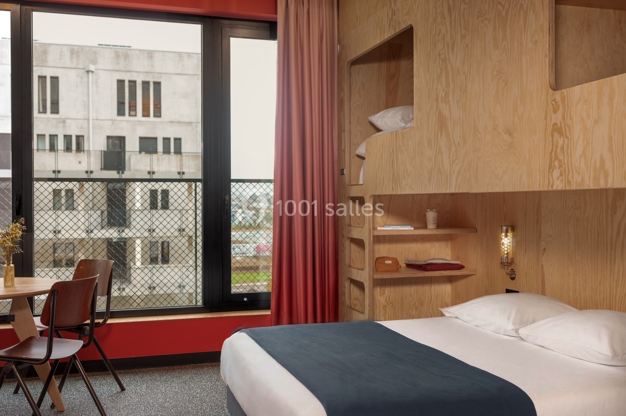 Chambre d'hôtel moderne avec lit double, étagères en bois, table et chaises, vue sur un bâtiment extérieur.